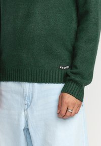Pull en tricot vert foncé avec des poignets et un ourlet côtelés, accompagné d'une petite étiquette noire "Volcom" sur le côté. Assorti à un jean bleu clair.