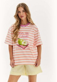 Mladá žena s dlhými vlnitými blond vlasmi má na sebe ružovo-biele pruhované tričko so zelenými jablkami a textom, v kombinácii s bledohnedými šortkami.