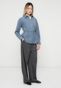 Vrouw die staat in een blauwe shirtjasje met riem, brede grijze broek en zwarte flats, tegen een effen lichte achtergrond.