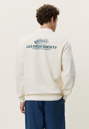 Homme aux cheveux bouclés porte un sweat crème avec l'inscription "LES DEUX SOCIETY PASSION FOR PEOPLE" et un motif globe à l'arrière, assorti à un pantalon bleu.
