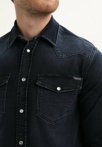 Donkerblauwe denim shirt met een western-stijl ontwerp. Voorzien van drukknoopsluitingen, dubbele borstzakken en een licht vervaagde textuur.