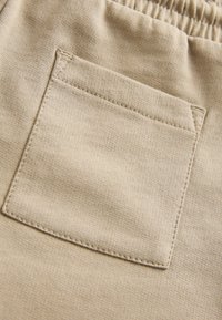 Béžová tkanina s hladkou texturou, na pravé straně má přišitou jedinou čtvercovou kapsu. Kapsa má obdélníkový tvar a viditelný šev.