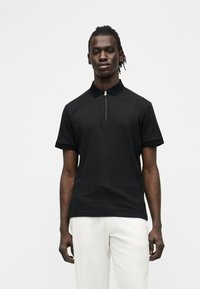 Homme debout, bras détendus, portant un polo noir à manches courtes avec fermeture éclair et un pantalon blanc, devant un fond clair uni.