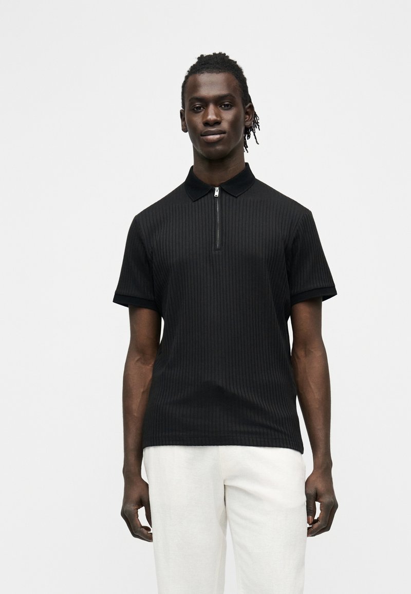 Homme debout, bras détendus, portant un polo noir à manches courtes avec fermeture éclair et un pantalon blanc, devant un fond clair uni.