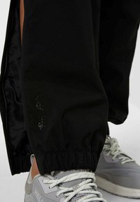 Pantalones impermeables negros con puños elásticos y aberturas laterales. Presentan la marca "HELLY TECH" en impresión reflectante. Combinados con zapatillas grises.