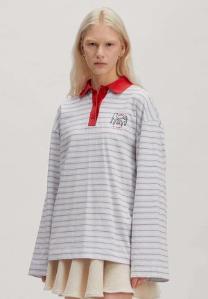 Jeune personne aux longs cheveux blonds porte un polo rayé ample avec un col rouge et un graphisme "Take a Seat", associé à une jupe beige.