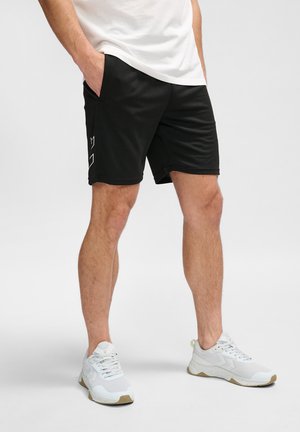 Mann trägt schwarze Shorts mit weißem geometrischem Logo auf der linken Seite, weiße Sneakers und ein weißes T-Shirt, steht mit den Händen in den Taschen.