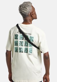 T-shirt color crema realizzato in cotone, con un design grafico sulla parte posteriore che presenta illustrazioni verdi in un motivo a griglia e un logo.