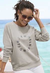 Helles graues Sweatshirt mit rundem Halsausschnitt und gerippten Bündchen, versehen mit einem kreisförmigen Textdesign in dunklerem Grau. Trägt dazu weiße Hosen.