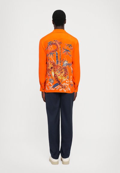 Helles oranges Hemd mit Tigermuster, lange Ärmel und geknöpften Manschetten. Kombiniert mit dunkelblauen Hosen und weißen Schuhen.