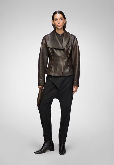 Brown leather motorcycle jacket with a wide collar, asymmetric zipper, and textured finish, paired with black tapered trousers and ankle boots.
