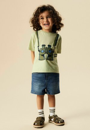 Niño sonriente con cabello rizado que lleva una camiseta verde claro con estampado de prismáticos, pantalones cortos de mezclilla, calcetines a rayas y sandalias marrones.