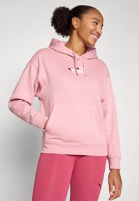 Lys rosa hettegenser med frontlomme, snøring i hetten og hvit logo. Myk stoff, avslappet passform, kombinert med matchende rosa tights.