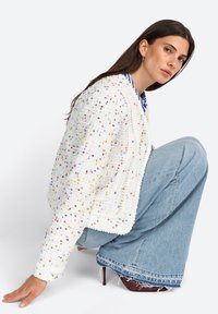 Weißer, strukturierter Blazer mit bunten Sprenkeln, kurzes Design und weichem Stoff. Kombiniert mit hellblauen, weit geschnittenen Jeans und dunklen Schuhen.