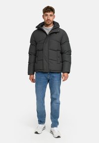 Man die staat in een zwart gewatteerd jack met capuchon, een grijze trui, blauwe jeans en witte sneakers tegen een effen lichte achtergrond.