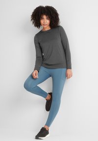Long-sleeve gray athletic top with a round neckline, paired with blue leggings and black sneakers, posing against a neutral background.