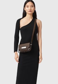 Ruskea nahkainen olkalaukku, jossa on läppä, hopeinen suorakulmainen solki ja irrotettava hihna, esiteltynä neutraalia taustaa vasten.