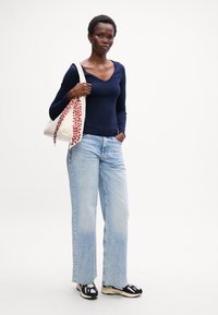 Cămașă albastru marin cu mânecă lungă, blugi largi albaștri deschiși și adidași negri și crem. Modelul ține o geantă tote cu o curea cu un model floral.