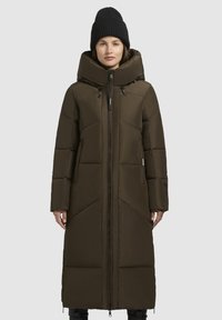 Dlhý hnedý prešívaný kabát s vysokým golierom, predným zipsom a veľkými vreckami. Má priliehajúcu siluetu a zväčšenú kapucňu. Čierny pletený čiapka.