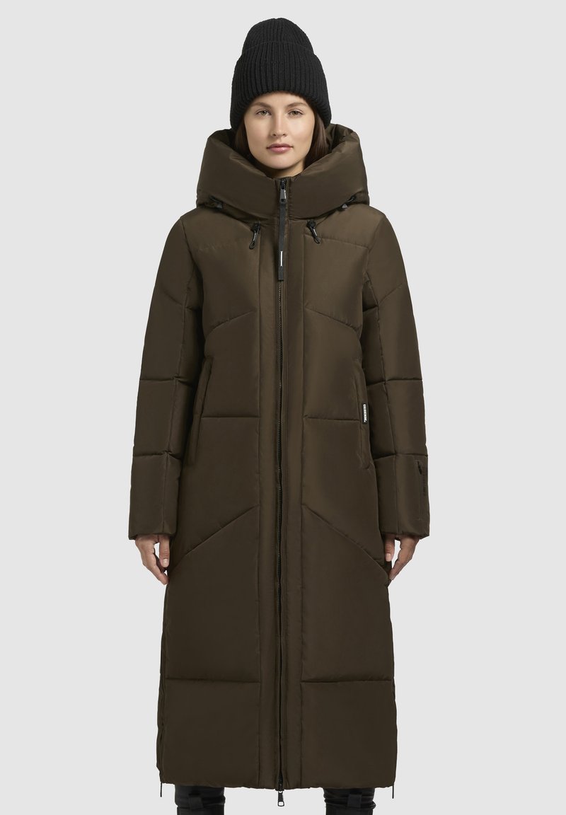 Dlhý hnedý prešívaný kabát s vysokým golierom, predným zipsom a veľkými vreckami. Má priliehajúcu siluetu a zväčšenú kapucňu. Čierny pletený čiapka.