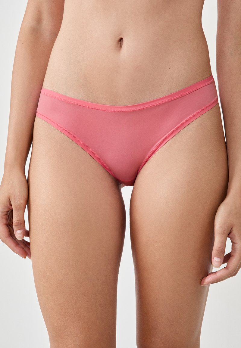 Donna che indossa mutande a forma di bikini rosa a vita bassa, inquadra dalla vita fino a metà coscia su uno sfondo neutro.