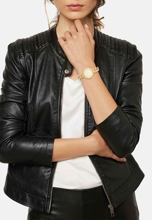 Kvinde iført en sort læderjakke og hvid skjorte, der viser et guldfarvet armbåndsur med ledkæde på venstre håndled.