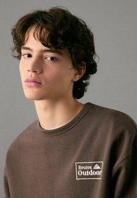 Bruine trui met een wit geborduurd "Routes Outdoor" logo. Ronde hals met geribbelde accenten en zachte, effen stoftextuur.