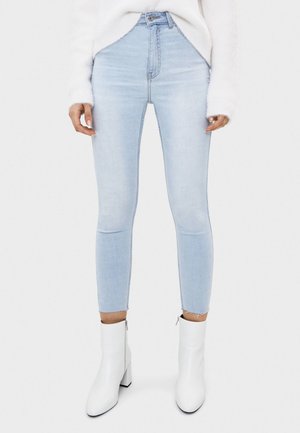 Femme portant un jean skinny bleu clair avec des ourlets effilochés, un pull blanc duveteux et des bottines blanches à talons.