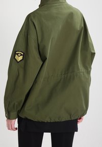 Oliwkowa kurtka z wysokim kołnierzem, elastycznym dołem i czarną haftowaną naszywką na lewym rękawie. Wykonana z trwałego, gładkiego materiału.