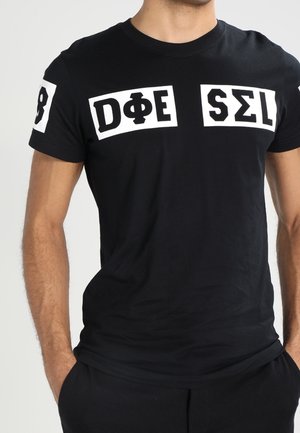 Mand iført sort t-shirt med hvide rektangulære mærker, der viser stiliserede bogstaver inklusive D, Φ, E, S, Σ og L på brystet og ærmet.