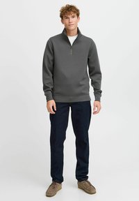 Dark gray zip-up sweatshirt with a high collar, long sleeves, and ribbed hem, paired with dark jeans and light brown shoes.