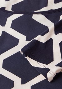 Tkanina w kolorze granatowym i kremowym z wzorem geometrycznym charakteryzuje się gładką teksturą. Posiada detal złożonego rękawa oraz widoczne szwy.