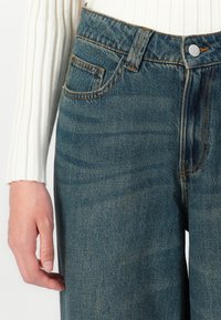 Mão esquerda descansando ao lado de jeans de denim azul com costura laranja e uma blusa de manga longa branca e canelada visível na cintura e no braço.