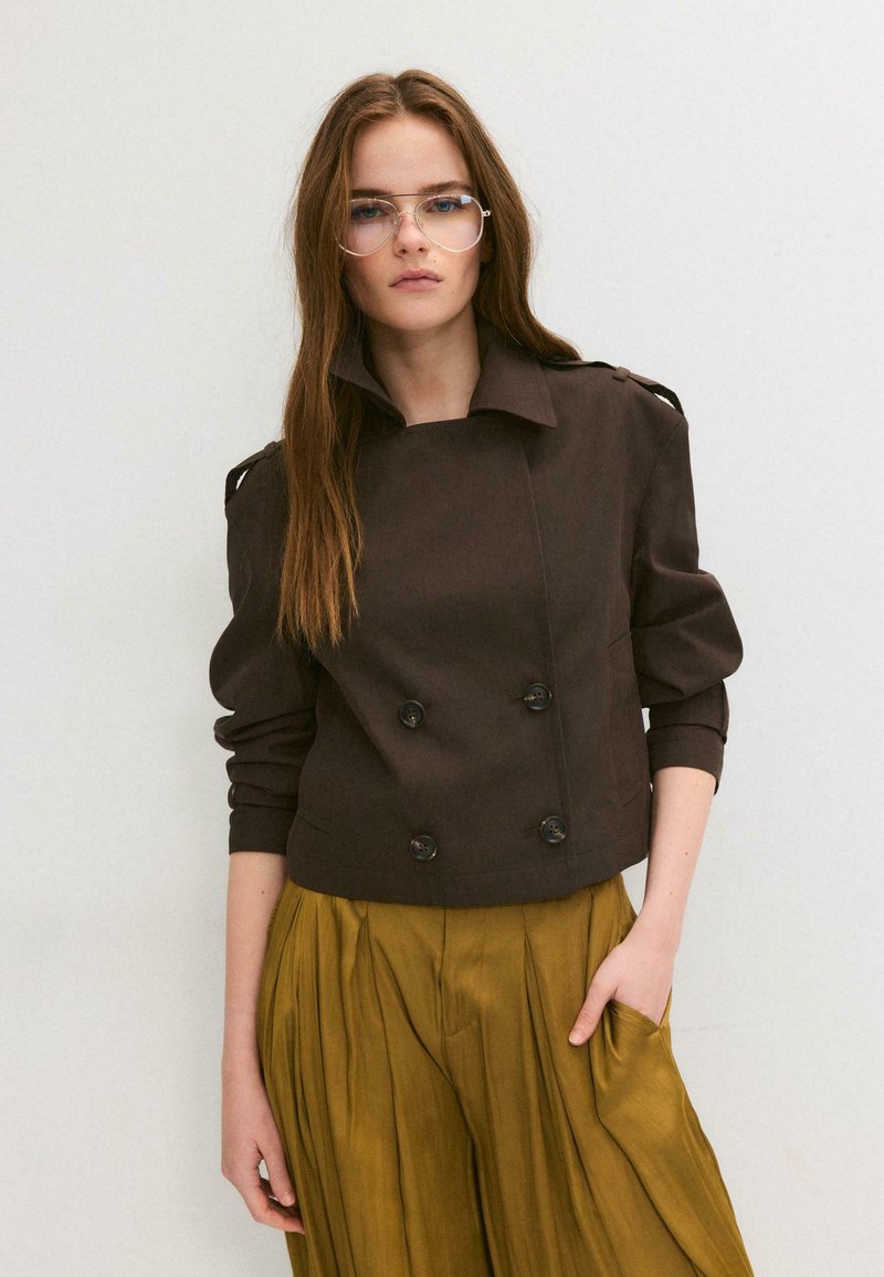 Young woman with long brown hair wearing clear round glasses, a dark brown double-breasted jacket, and mustard yellow pleated pants.