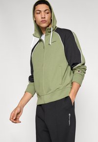 Sweat à capuche zippé vert et noir avec des accents blancs, doté d'une capuche, de poches kangourou et d'un logo brodé sur la poitrine et la manche.