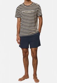 Beige- ja laivastonsinisiksi raidallinen puuvillainen T-paita pyöreällä pääntiellä; yhdistetty laivastonsinisiin shortseihin. Malli seisoo paljain jaloin, kädet taskuissa.