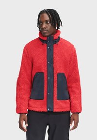Czerwona polarowa kurtka z granatowymi nylonowymi akcentami, z wysokim kołnierzem, zatrzaskami oraz dwoma przednimi kieszeniami, teksturowana dla zapewnienia ciepła.