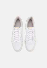 Zapatillas deportivas de cuero blanco con puntera redonda, perforaciones en la caja del dedo y detalles texturizados. Presentan cordones blancos y un logo dorado en la lengüeta.