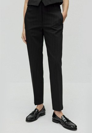 Zwarte op maat gemaakte broek met witte streepjes, slim fit, met omslag aan de onderkant. Draagt een paar glanzende zwarte loafers die een soepele leren textuur laten zien.