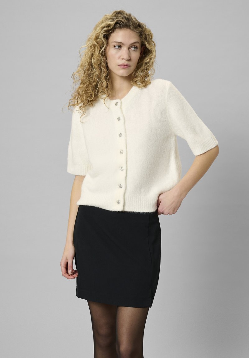 Valge lühikeste varrukatega kardigan pehmest, tekstureeritud kangast, mille esiosas on viis dekoratiivset nuppu, koos musta mini-seelikuga.