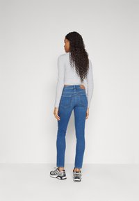 Jeans ajustados de color azul con bolsillos traseros, costuras de contraste y una etiqueta de cuero con marca. Combinados con una camiseta gris de manga larga y zapatos deportivos.
