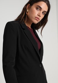 Sort skræddersyet blazer med en enkelt knap, klapper og en glat tekstur. Båret over en mørk burgunder top, der viser et professionelt look.