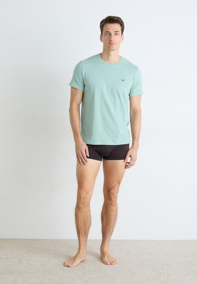 Un homme pieds nus portant un t-shirt vert clair et un slip noir devant un fond de mur blanc uni.