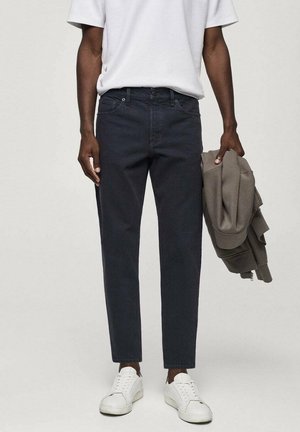 Jeans blu scuro con taglio dritto, indossati con una maglietta bianca e scarpe da ginnastica bianche, accompagnati da una giacca grigia tenuta in una mano.