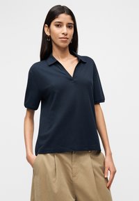 Μπλε σκούρο πόλο με κοντά μανίκια, λαιμόκοψη V και άνετη εφαρμογή. Το ύφασμα φαίνεται λείο με διακριτή υφή. Φοράται με χακί παντελόνια.