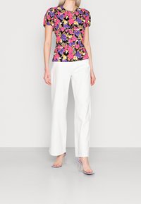 Il top floreale con maniche a sbuffo presenta fiori vibranti in rosa, viola e giallo su uno sfondo nero, abbinato a pantaloni bianchi a gamba larga e tacchi trasparenti.