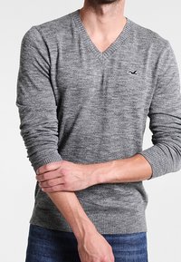 Šedý pletený svetr s výstřihem do V a žebrovanými manžetami. Obsahuje malé logo na hrudi a texturovaný vzor látky.