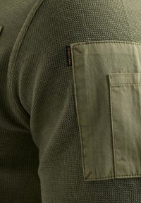 Olivgrüner, strukturiertes Strick-Sweatshirt mit einer flachen Tasche am Ärmel und einem kleinen Etikett. Der Stoff ist mit einem Rautenmuster gewebt.