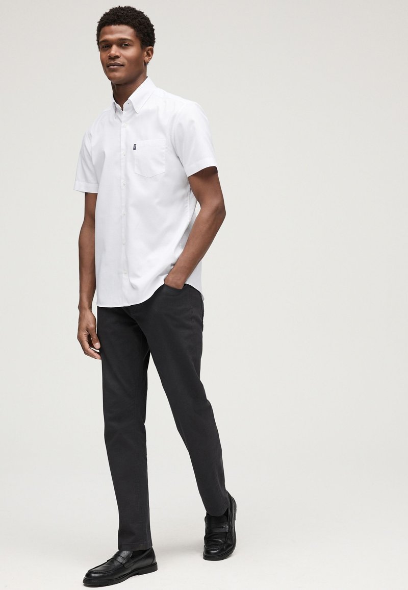 Homme debout, une main dans la poche, portant une chemise blanche à manches courtes boutonnée, un pantalon noir et des chaussures noires sur un fond uni.