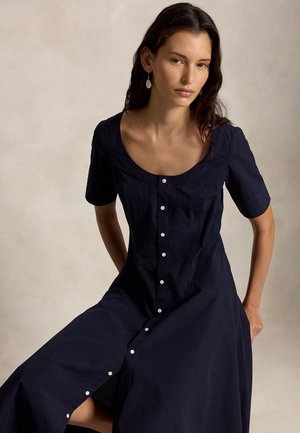 Žena s dlhými tmavými vlasmi má na sebe námornícky modré košeľové šaty s krátkymi rukávmi a bielymi gombíkmi, pózujúca na svetlom jednofarebnom pozadí.